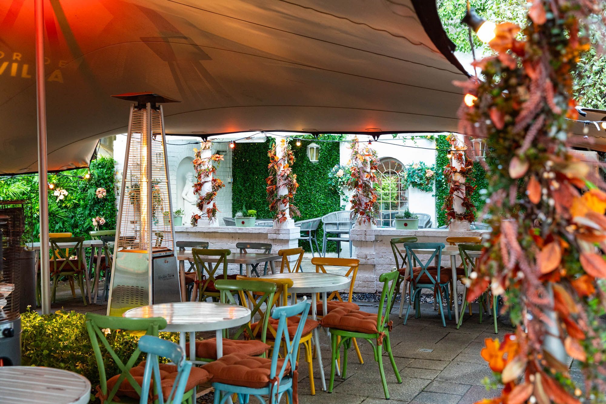 garden shot of Hove Place. Different colored chairs, white tables and umbrellas with outdoor heaters next to them
