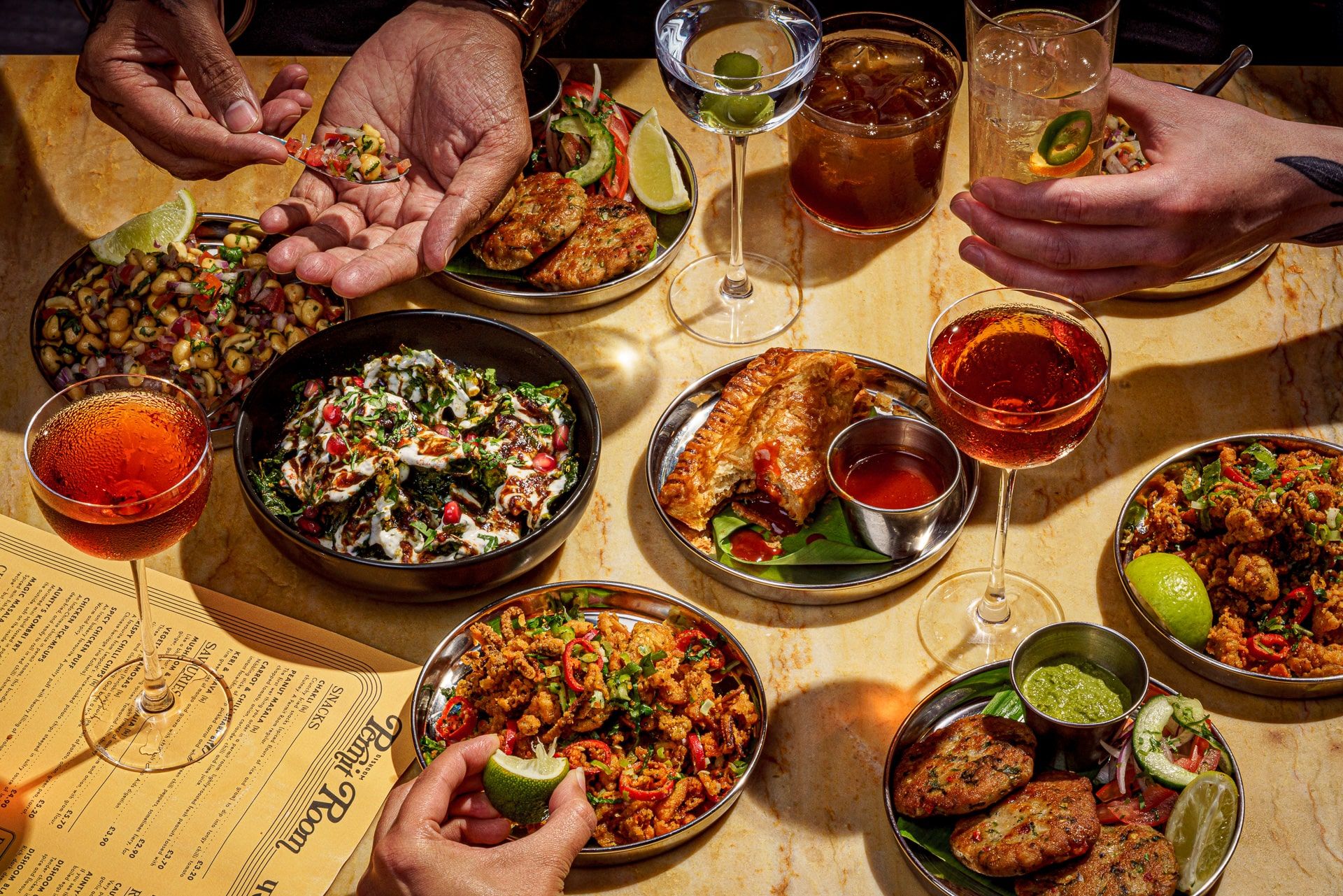 Lots of plates of food with people's hands in the shot eating the food. With drinks on the table to accompany the small plates of food. Photo Credit: Permit Room