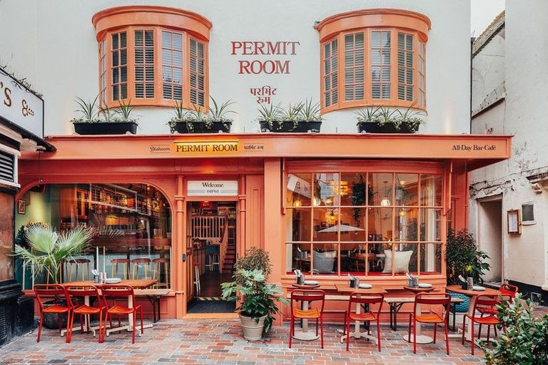 exterior shot of the beautiful Permit Room building, orange and white walls, with plant decorations, red chairs and orange tables.