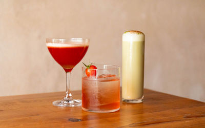 Three cocktails in different glasses on a wooden table with a tomato garnish.