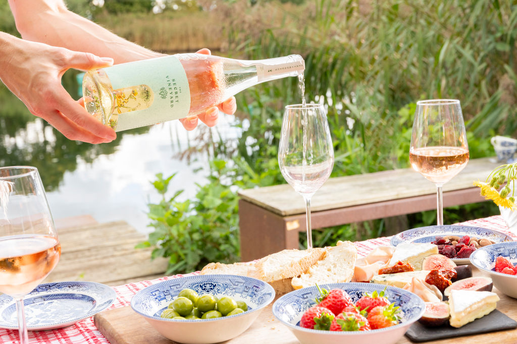 Wine being poured into a glass next to a lake with fresh fruit and a bowl of olives.