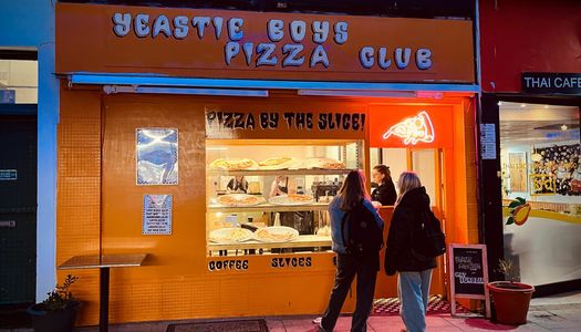 2 women stand outside an orange pizza shopfront that has pizzas in the window. New restaurants in Brighton in 2024