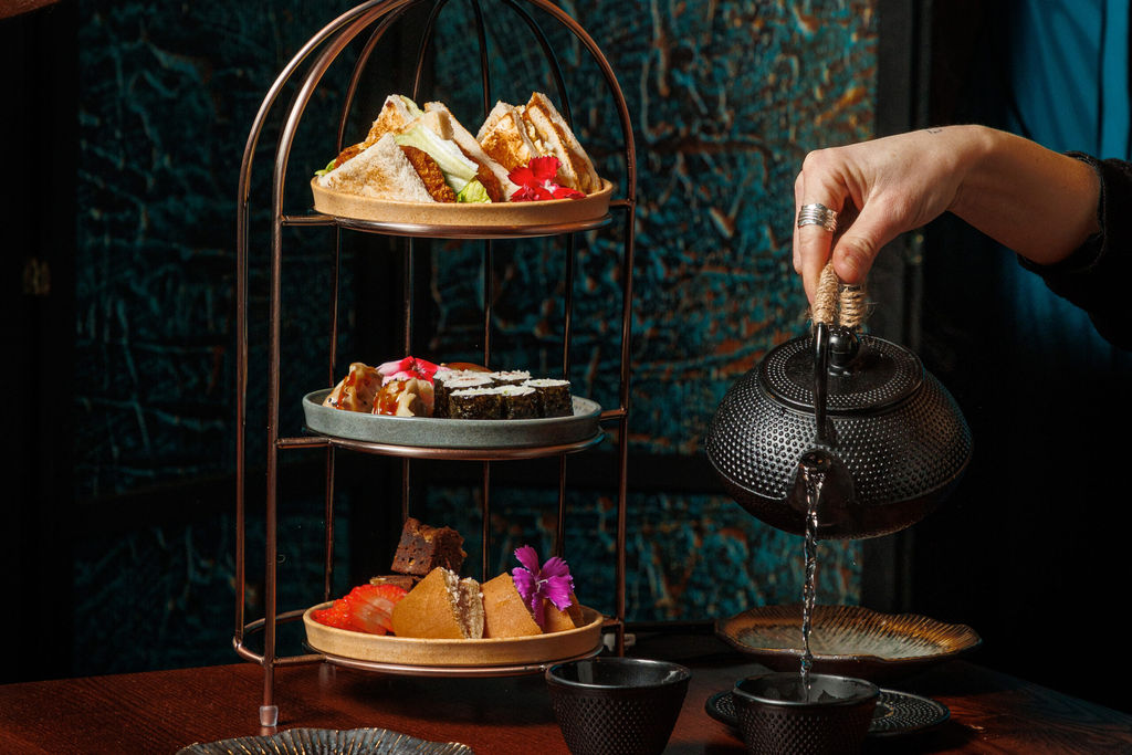 A tierred platter of savoury and sweet treats and a hand pouring a pot of tea.
