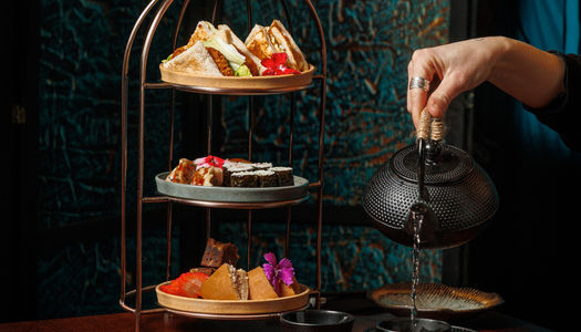A tierred platter of savoury and sweet treats and a hand pouring a pot of tea.