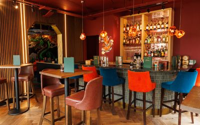 Tall tables with tall chairs inside an ambient lit room with a bar at the back sticked with various bottle of drinks.