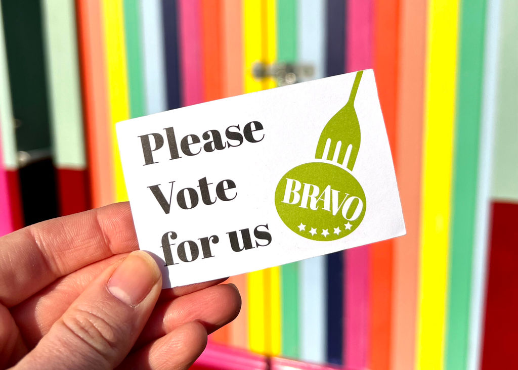 A BRAVO card being held up against rainbow coloured beach huts