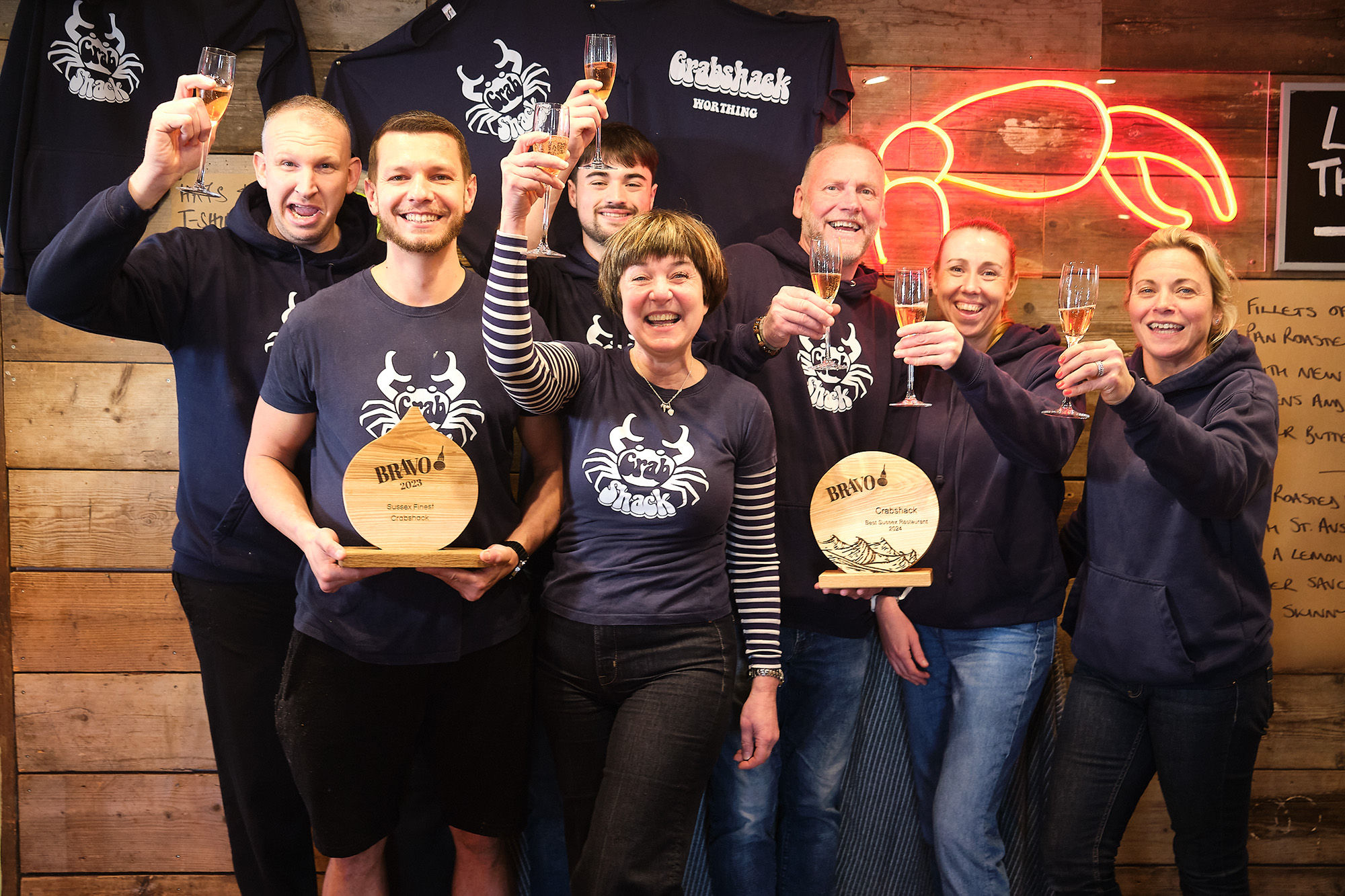 Crabshack team in Worthing. Their members of staff in branded T-Shirts against a wooden wall backdrop and a neon crab sign. Winner od the best Sussex restaurant in 2024.