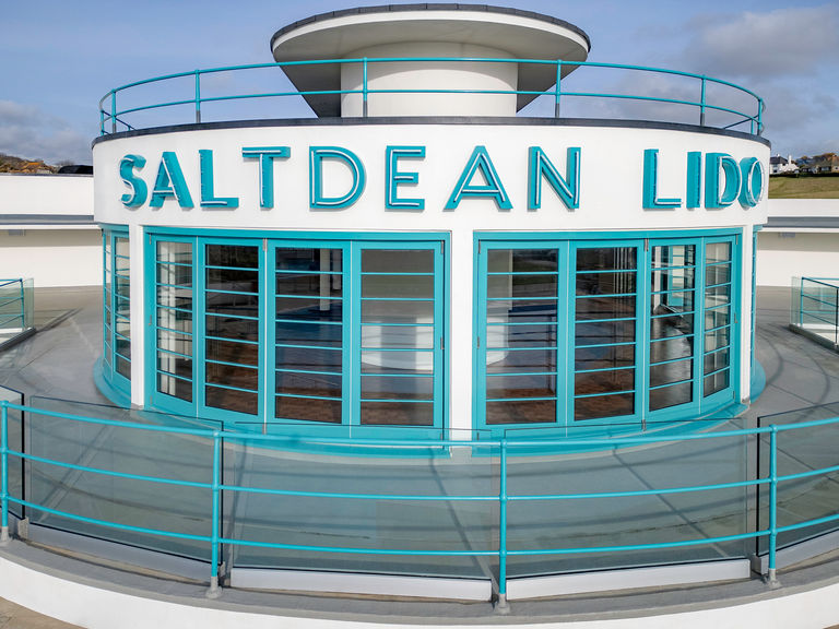 The first floor cyclindrical Deco restaurant facade at Saltdean Lido. White exterior with bug curved windows and blue trim on railings and windows.