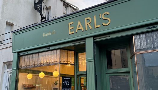 Earls sandwich shop with sandwich shop signage in gold, against a green facade. A warm glow coming the window against a backdrop of a blue and cloudy sky.