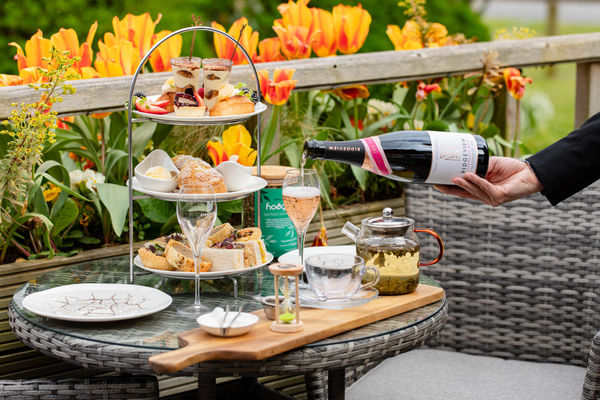 An afternoon tea set up on a table outside with orange tulips in the background and a glass of sparkling wine being poured.