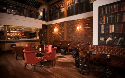 A dark and inviting hotel bar with leather chairs and dark walls. Chandelier lighting hangs overhead. The feel is an old gentlemans club.