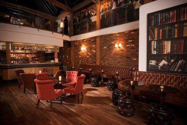 A dark and inviting hotel bar with leather chairs and dark walls. Chandelier lighting hangs overhead. The feel is an old gentlemans club. 