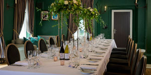 A dark green dining room is elegantly decorated for a wedding. A long banquet table has a white tablecloth and is dressed for a wedding. Crokery, candlesticks and glasses. It looks very classically beautiful and warm.