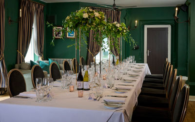 A dark green dining room is elegantly decorated for a wedding. A long banquet table has a white tablecloth and is dressed for a wedding. Crokery, candlesticks and glasses. It looks very classically beautiful and warm.
