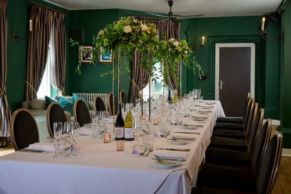 A dark green dining room is elegantly decorated for a wedding. A long banquet table has a white tablecloth and is dressed for a wedding. Crokery, candlesticks and glasses. It looks very classically beautiful and warm. 