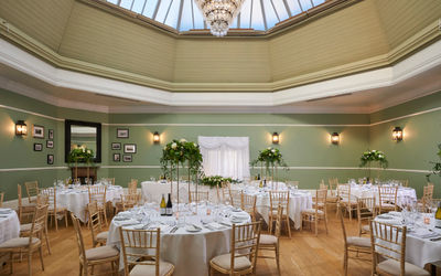 A large room adorned wiht a big chandelier is bright and elegant. There are round tables that are set up for a wedding, with white tablescloths and pretty chairs. The walls are a sage green with a cream rotunda that leads to a beautiful glass dome that lets in all the daylight.