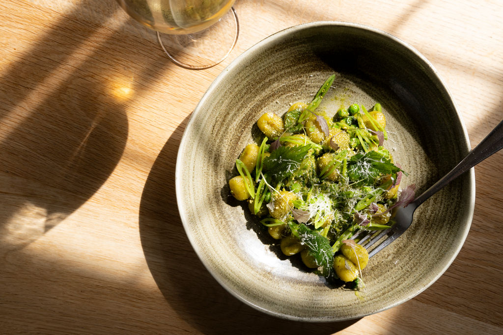 An overhead photo of a gnocchi dish with fresh greens and reflections from the sunlight shining against the bowl and glass of wine.
