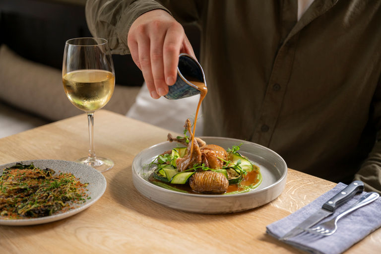 Sauce being poured pver a plate of food with a roasted quail and a glass of white wine next to the plate.