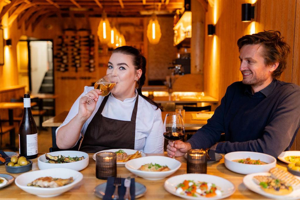 Head chef and owner of restaurant at a table with plates of food and drinking wine.