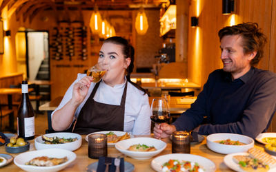 Head chef and owner of restaurant at a table with plates of food and drinking wine.