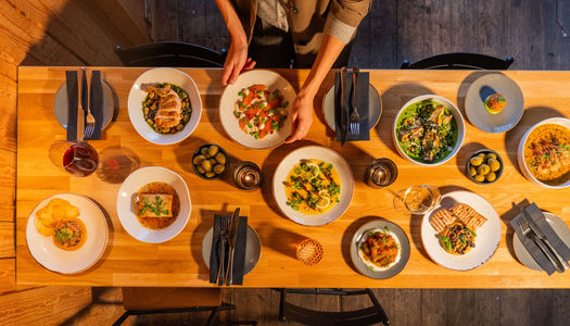 Overhead wide photo of a table with lots of different dishes of food all laid out and a man placing the final dish down.