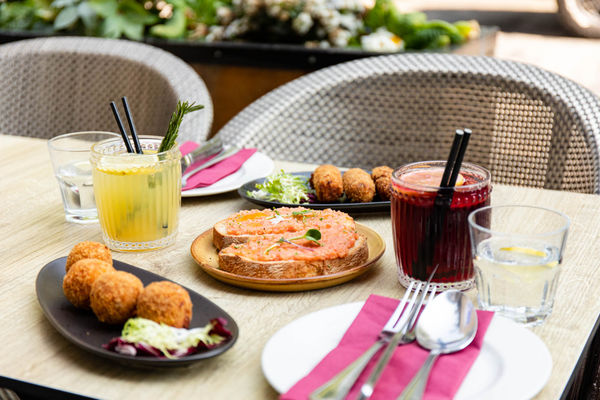 An alfresco dining set up. On the table is some spanish tapas and 2 cocktails. The table is set with cutlery and wine coloured napkin. The chairs are wicker.