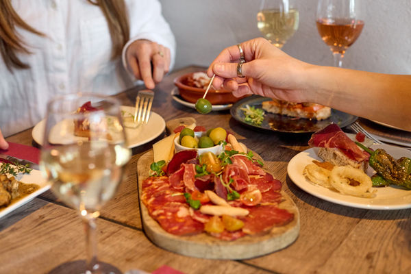 Two diners are at a table and sharing a spanish tapas platters. They are spearing olives, cutting cheeses. There is wine on the table as well as other small tapas plates. 