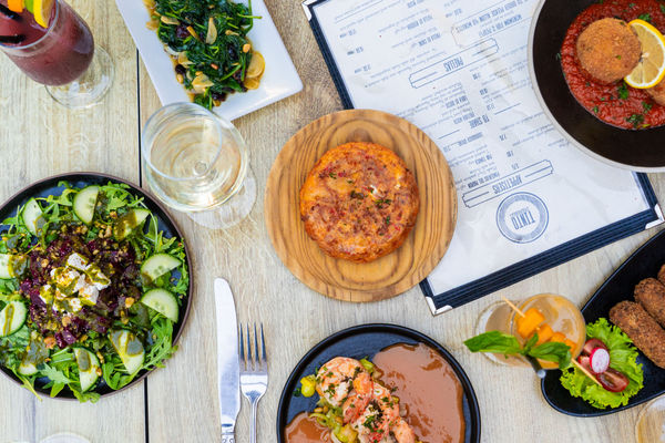 a sunny light coloured dining table. On top is a tapas menu and several small plates of Spanish Tapas plus a drink. The food looks fresh and tasty. 