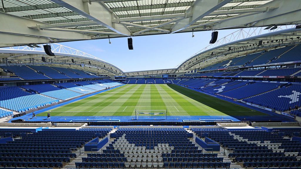 Pictured, the Brighton and Hove Football Stadium. a wide angle view which shows the stadium from inside. a beautiful lush green football pitch. the blue and white colours of the BHAFC or The Seagulls flag. Against the backdrop of a blue sky. The American Express Stadium is a venue where you can watch all the premiership games in their Sports Bar which is called Fanzone.