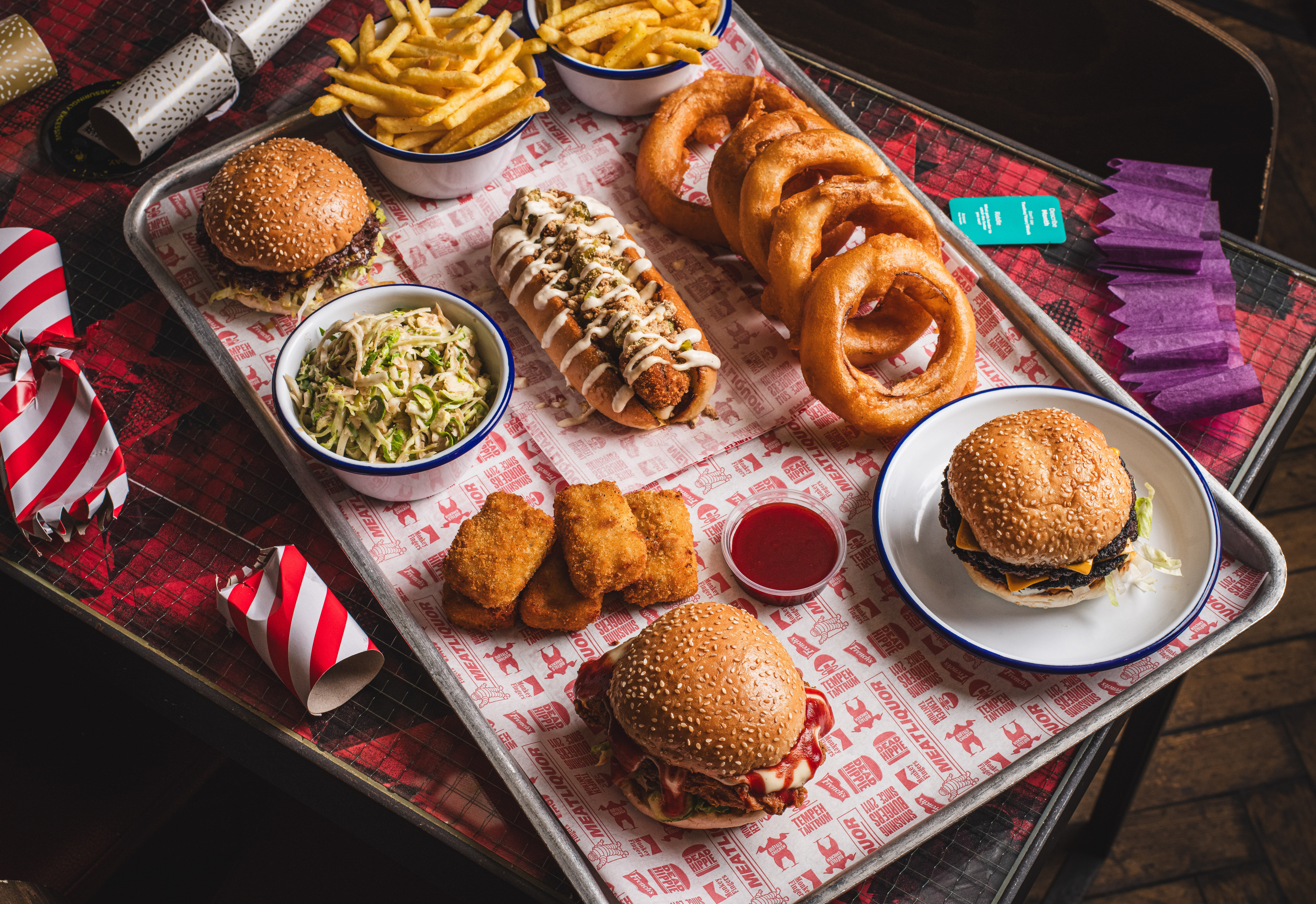 A tray with onion rings, a hot dog, burgers, fries and sides with festive props around the edge.