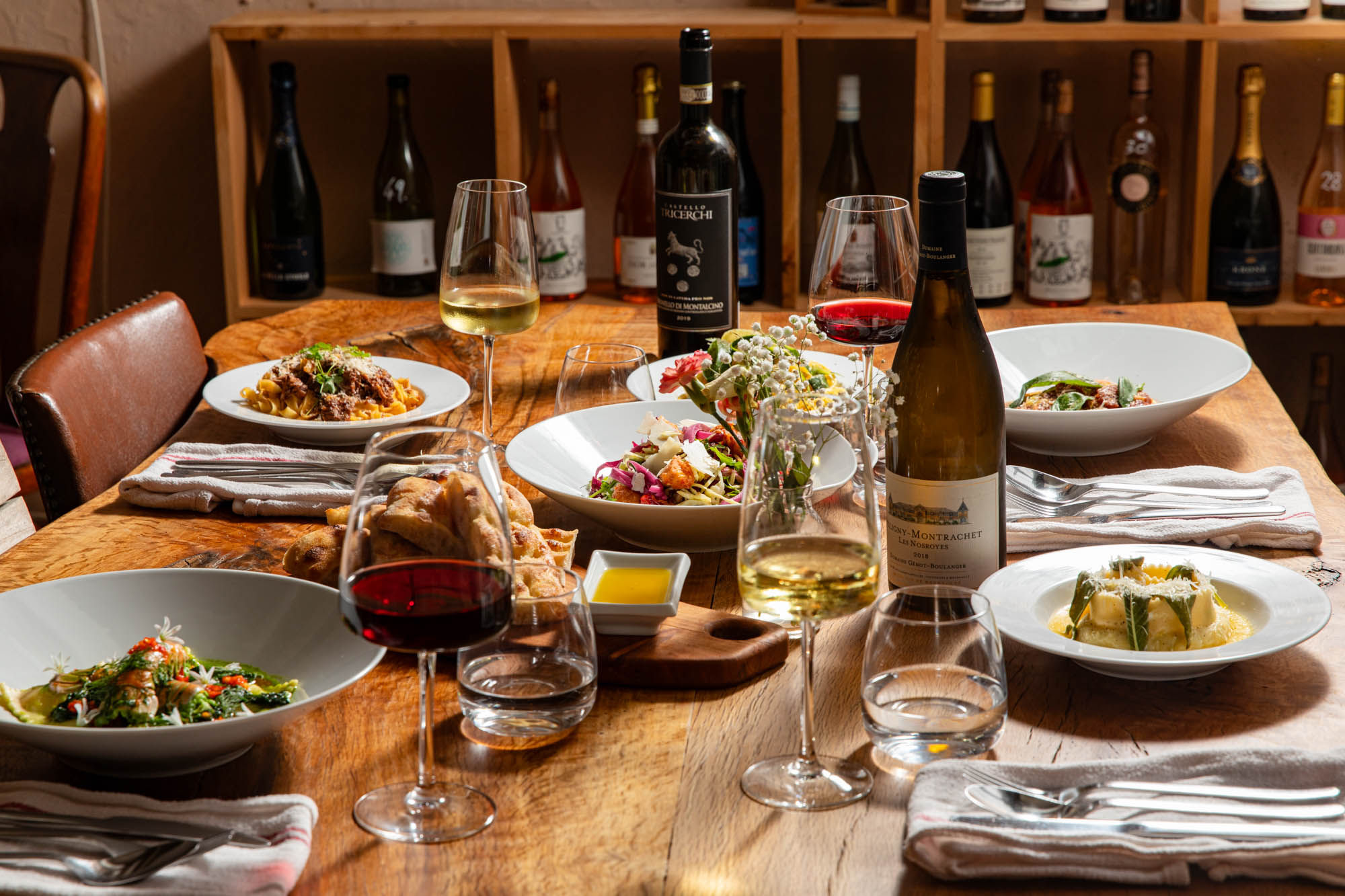 table laid out for a group of people. It includes multipe dishes with bottles of wine on the table