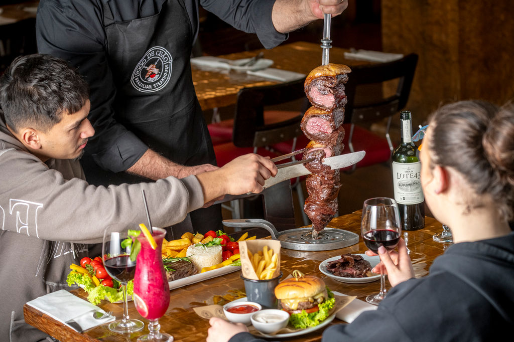 a man cutting of the meat of the stick at Fogo de Chão Brazilian Steakhouse. Enjoy an all-you-can-eat churrasco steak experience, with tableside-carved signature cuts like grilled Picanha, pork loin, lamb, chicken sausage, and ribs.