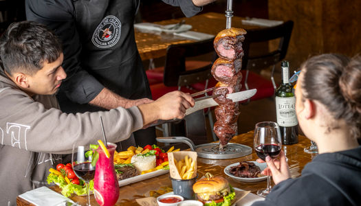 a man cutting of the meat of the stick at Fogo de Chão Brazilian Steakhouse. Enjoy an all-you-can-eat churrasco steak experience, with tableside-carved signature cuts like grilled Picanha, pork loin, lamb, chicken sausage, and ribs.