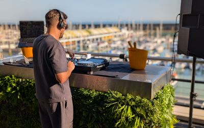 A Dj is playing on the baacony of a hotel that overlooks a marina. Lots of boats can be seen behind the DJ as he mixes music on the decks.
