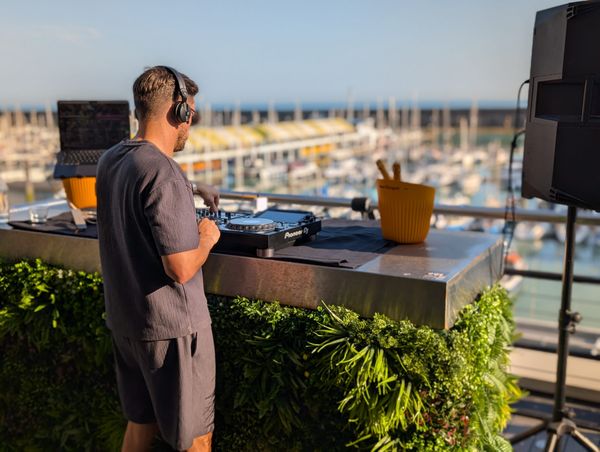 A Dj is playing on the baacony of a hotel that overlooks a marina. Lots of boats can be seen behind the DJ as he mixes music on the decks. 