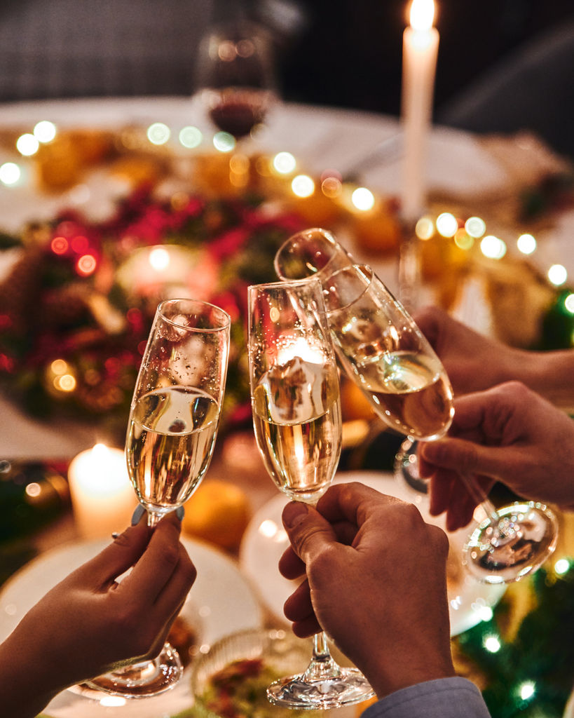A group of people holding champagne glasses with fairy lights and christmas food int he background.