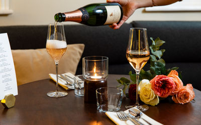 Two glasses of sparkling wine being poured, sat on a table with a tealight candle in the middle and a bunch of pink flowers on the table.