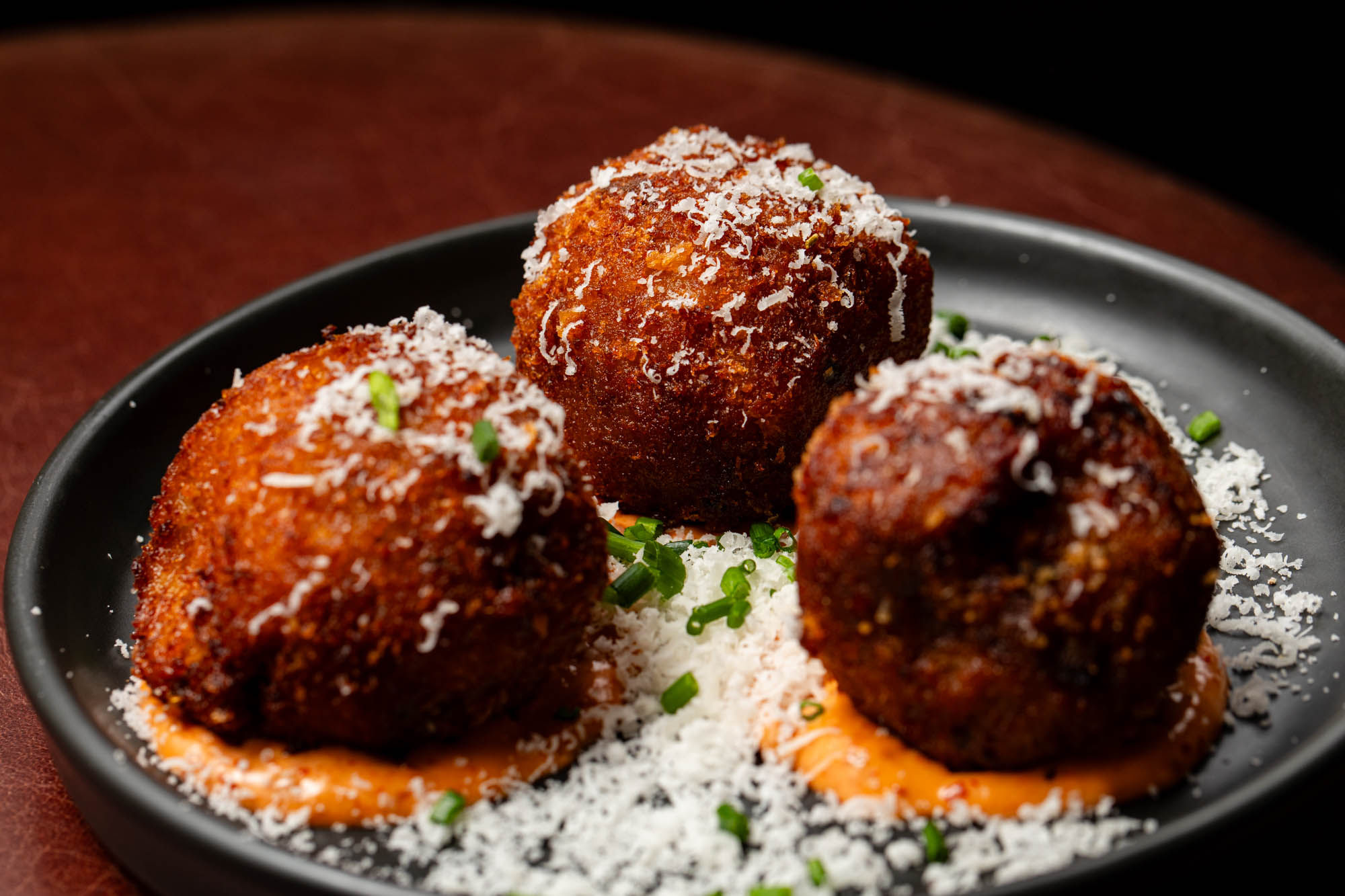 Three golden, crispy arancini balls served on a black plate, resting on a bed of creamy orange sauce, garnished with grated cheese and finely chopped chives.