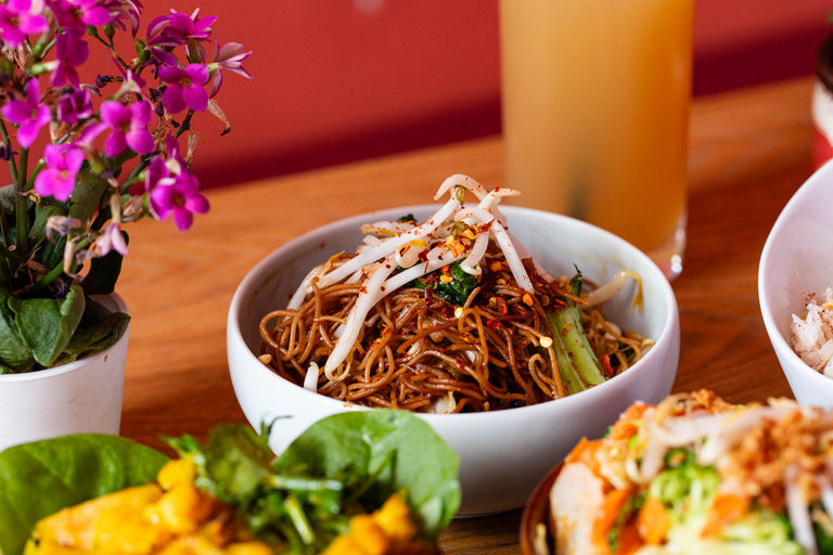 A bowl of noodles in a bowl with bean sprouts on top next to a pot with a pink plant.