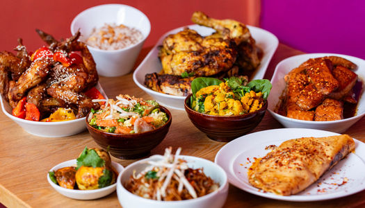 A variety of Asian dishes displayed on a wooden table with a bright pink wall behind.