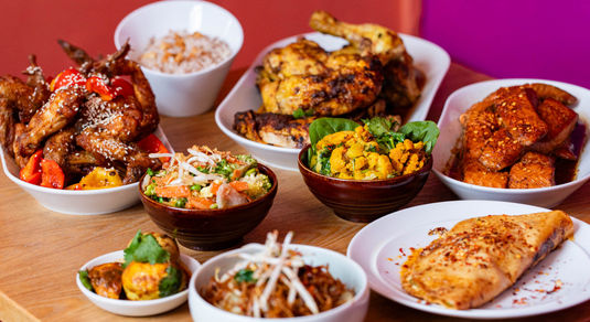 A variety of Asian dishes displayed on a wooden table with a bright pink wall behind.