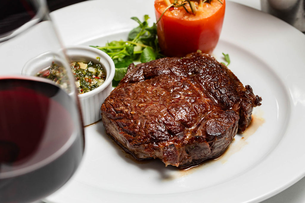 A perfectly seared steak at Il Bistro, served with a side of fresh chimichurri, a roasted tomato, and a garnish of greens. The dish is paired with a glass of rich red wine, making for a classic and indulgent dining experience in the heart of Brighton.