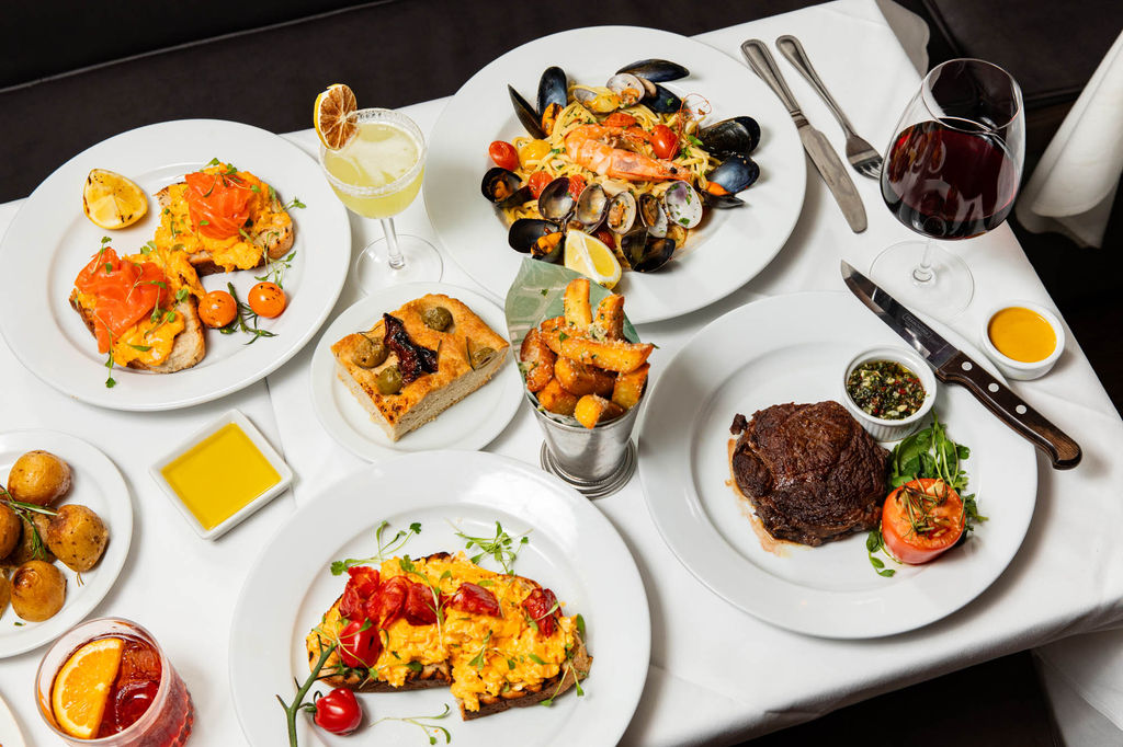 A beautifully arranged spread of Il Bistro’s finest dishes, featuring fresh seafood pasta with mussels and prawns, a perfectly seared steak with chimichurri, and vibrant scrambled eggs on toast with smoked salmon and cherry tomatoes. The table is adorned with crispy parmesan fries, warm focaccia, and golden roasted potatoes, accompanied by rich red wine, a citrusy cocktail, and a classic Negroni—an elegant feast capturing the essence of classic European dining.