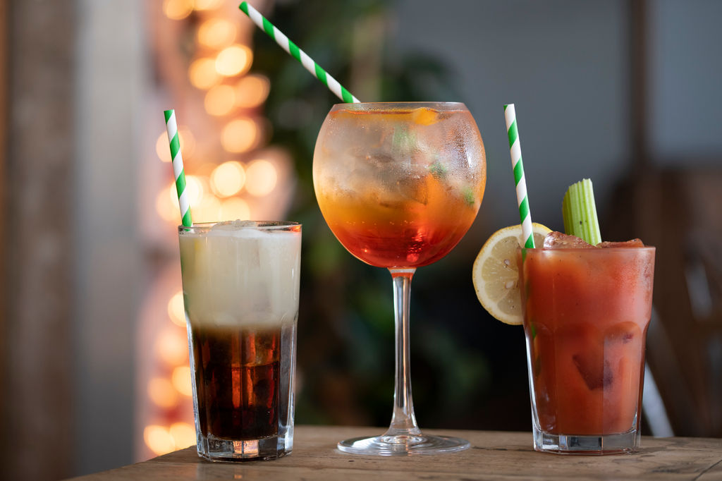 A selection of happy hour cocktails from a Brighton bar. Pictured an Aperol Spritz centre, a bloody Mary and an unknown cocktail. The 3 cocktails are super colourful with twinkly lights in the background.