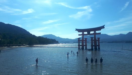 This image captures the stunning floating torii gate of Itsukushima Shrine on Miyajima Island, Japan, standing gracefully in the shallow waters as visitors walk around it. The scene is bathed in sunlight, reflecting off the water’s surface, with a backdrop of lush green mountains and a clear blue sky. A group of people can be seen wading through the shallow tide, adding a sense of scale to this iconic cultural landmark.