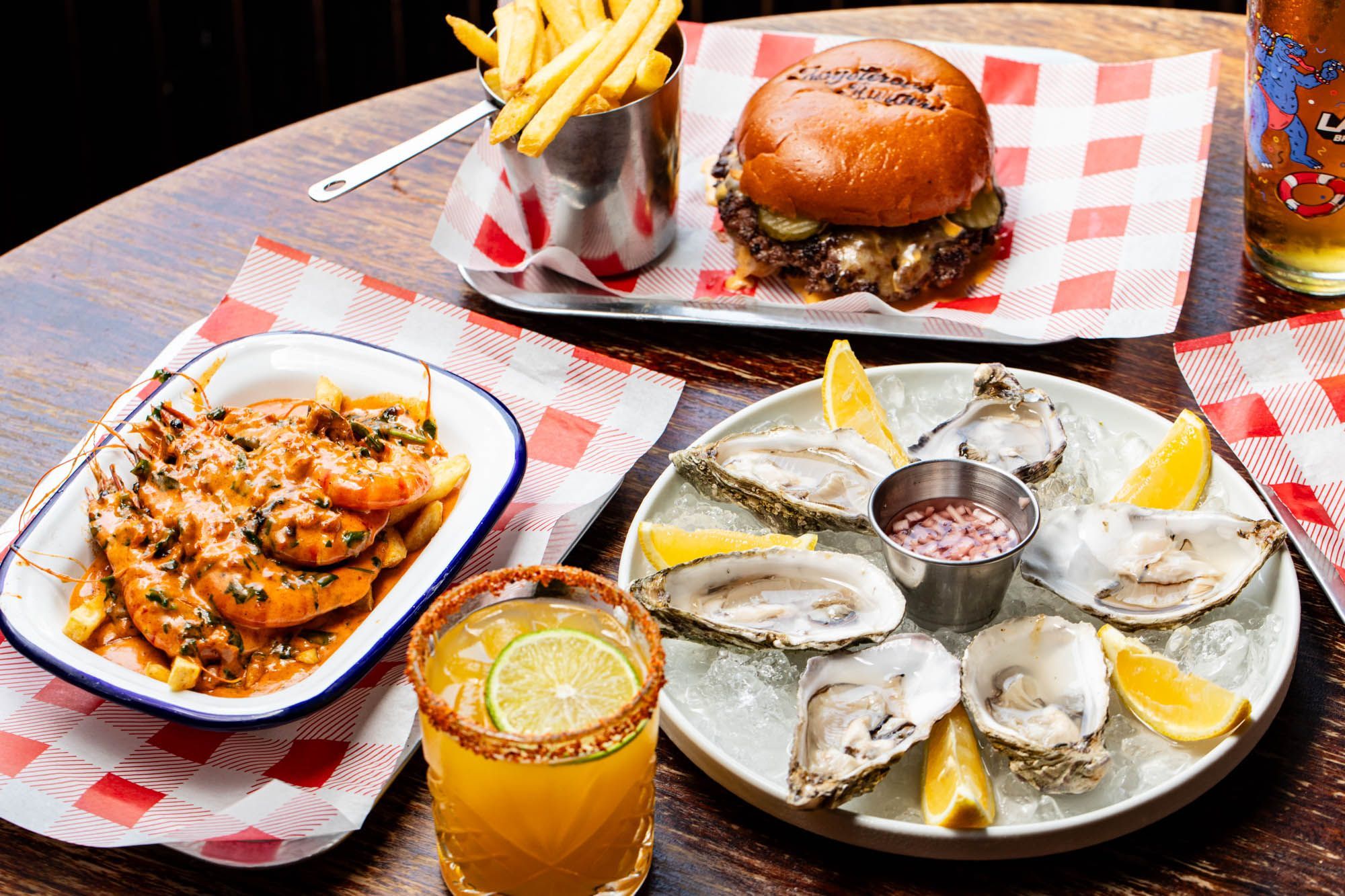 side shot of the wooden table with burger and fries, oysters, shrimps and cocktails at Boysterous Burgers