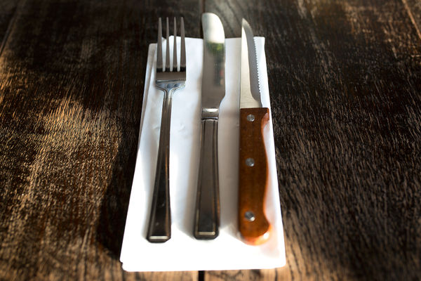 A knife and fork, with a steak knife on a serviette on a wooden table. This image showcases our best restaurants in Brighton page at the 2025 Brighton Restaurant Awards. This page showcases teh public voted best restaurants or top 20 restaurant in Brighton and Hove.
