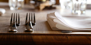 a picture of a place setting at a restaurant with two forks on the left hand side and a linen napkin on the wooden table. Glasses are sparkling in the background. This image represents the Restaurants Brighton Gift Vouchers page where gift vouchers can be redeemed across multiple locations in Brighton, Hove and Sussex.