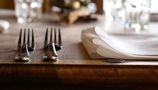a picture of a place setting at a restaurant with two forks on the left hand side and a linen napkin on the wooden table. Glasses are sparkling in the background. This image represents the Restaurants Brighton Gift Vouchers page where gift vouchers can be redeemed across multiple locations in Brighton, Hove and Sussex.