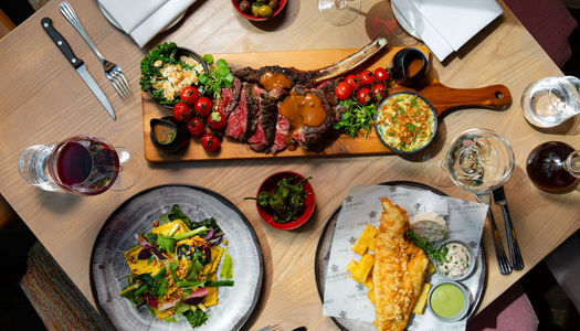 A table full of delicious food. One large steak platter in the centre with sauces and garnish. A fish and chips and a pasta dish. The table is set and there are glasses of wine.